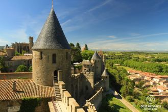 The walls of Carcassonne and its medieval towers from above. Blue skies, small houses on the right, green trees and snowy mountains in the background.