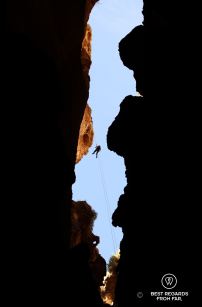 One person free abseiling down a rope to explore the Seventh Hole Cave in Oman. The abseil is so long (120 meters) that the caver looks tiny against the blue sky.
