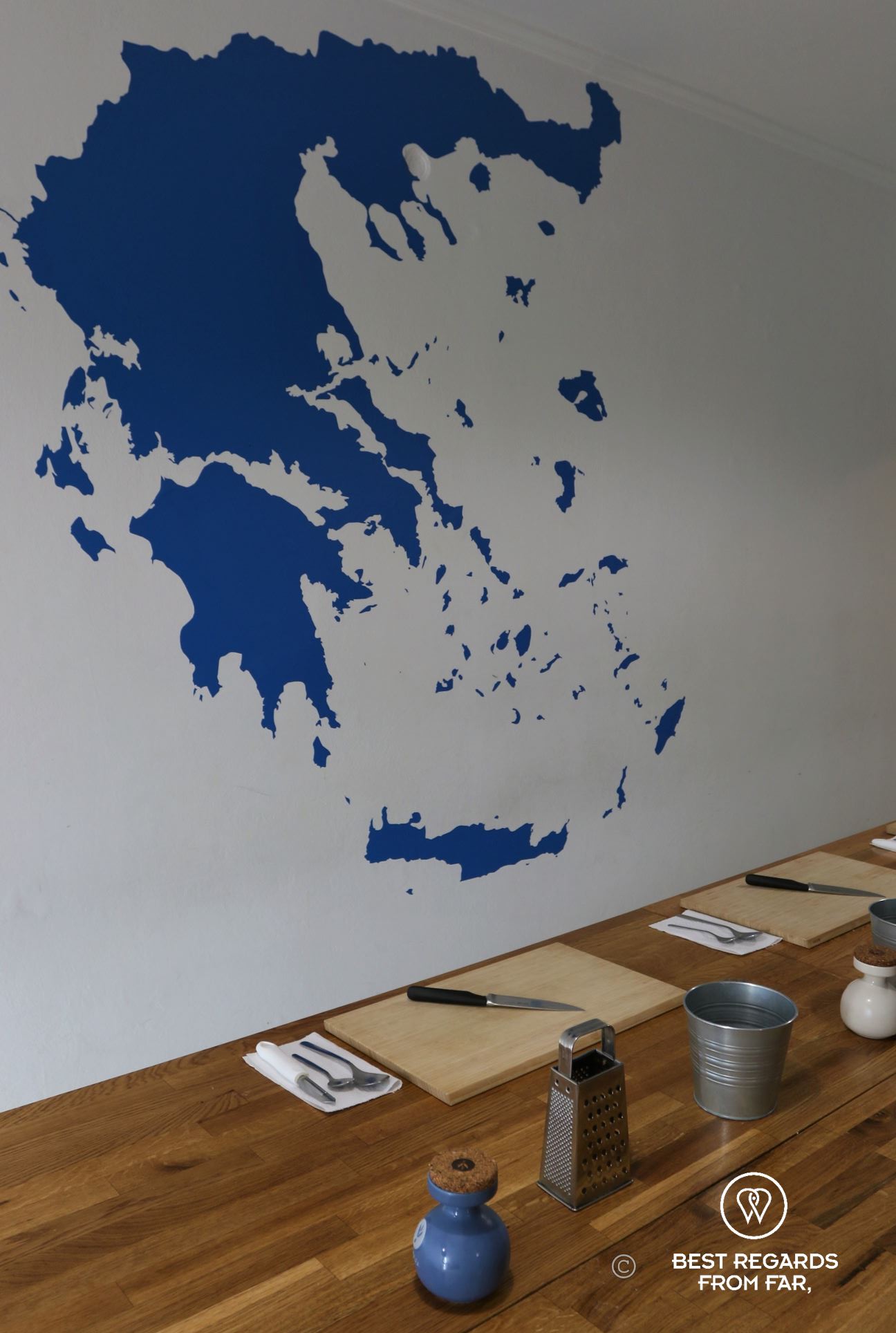 Greece painted in blue on a white wall and wooden table with chopping boards and cutlery.